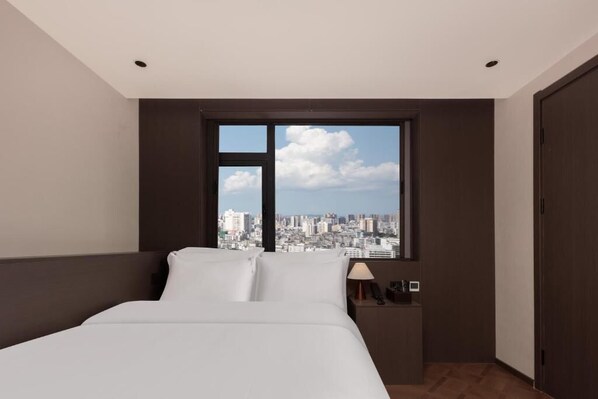 A hotel room with a bed, a nightstand, a lamp, and a window with a city view.