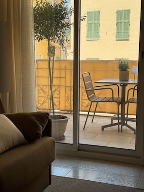 Una habitación con sofá, planta en maceta, mesa con sillas y vista a un edificio amarillo con persianas verdes a través de una puerta de vidrio.