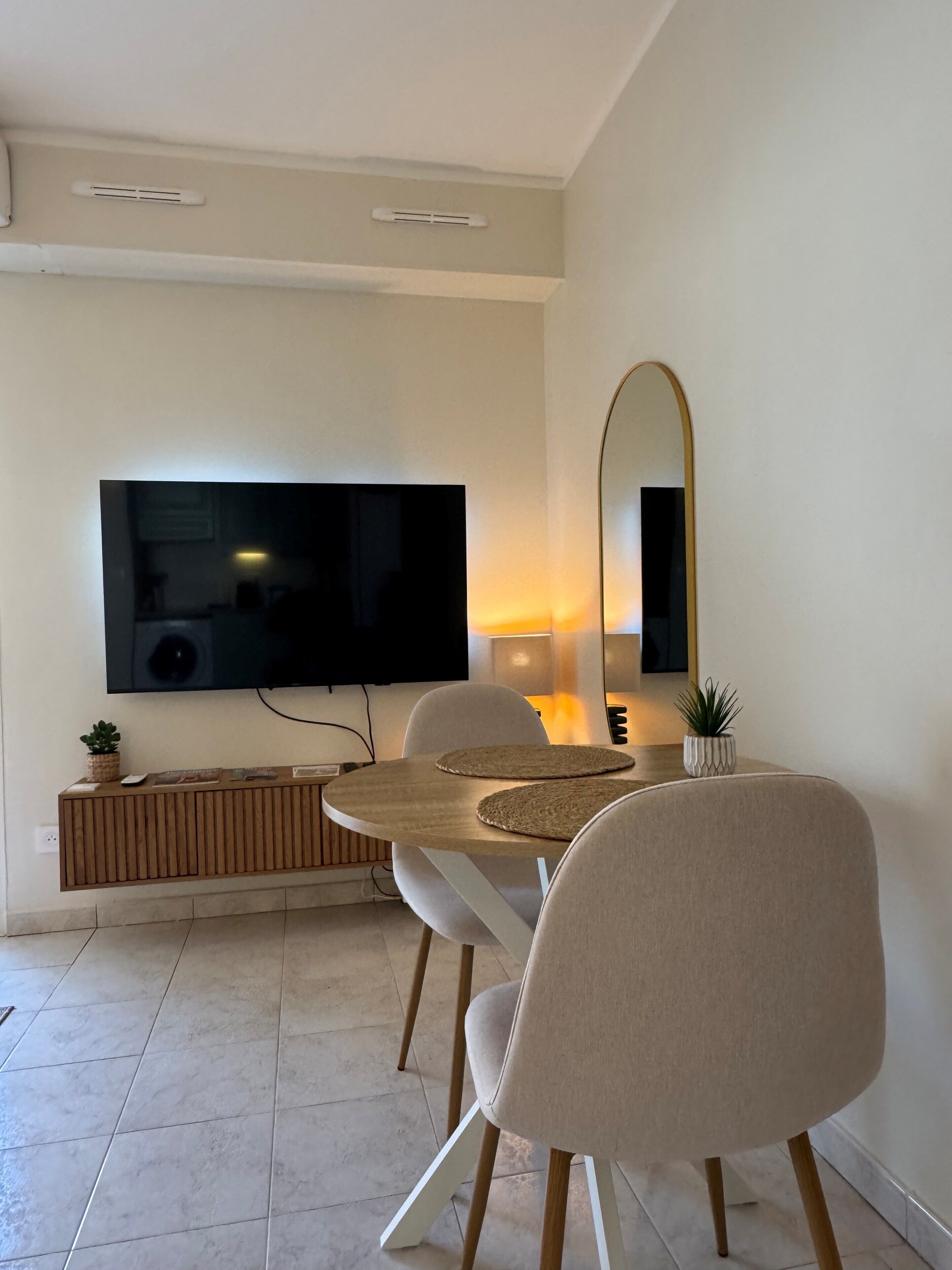 Un salón moderno con un televisor de pantalla plana montado en la pared, una mesa de comedor redonda de madera con dos sillas, una planta pequeña en maceta y un espejo.