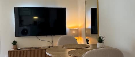 Un salón moderno con un televisor de pantalla plana montado en la pared, una mesa de comedor redonda de madera con dos sillas, una planta pequeña en maceta y un espejo.