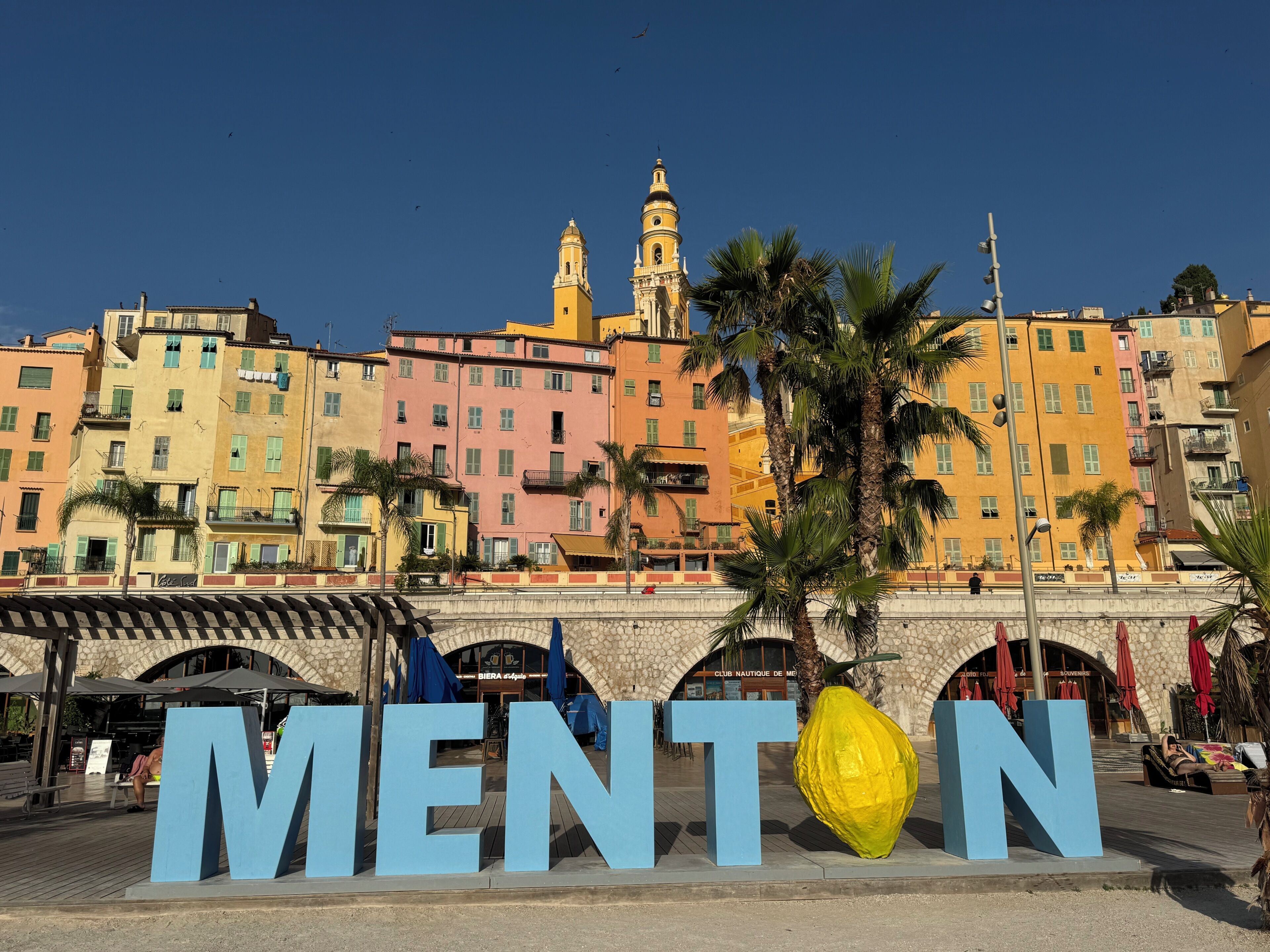 Une ville côtière aux bâtiments aux couleurs vives, avec une grande enseigne portant l’inscription « MENTON » et un pont en pierre.