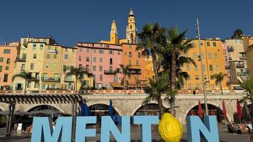 Une ville côtière aux bâtiments aux couleurs vives, avec une grande enseigne portant l’inscription « MENTON » et un pont en pierre.