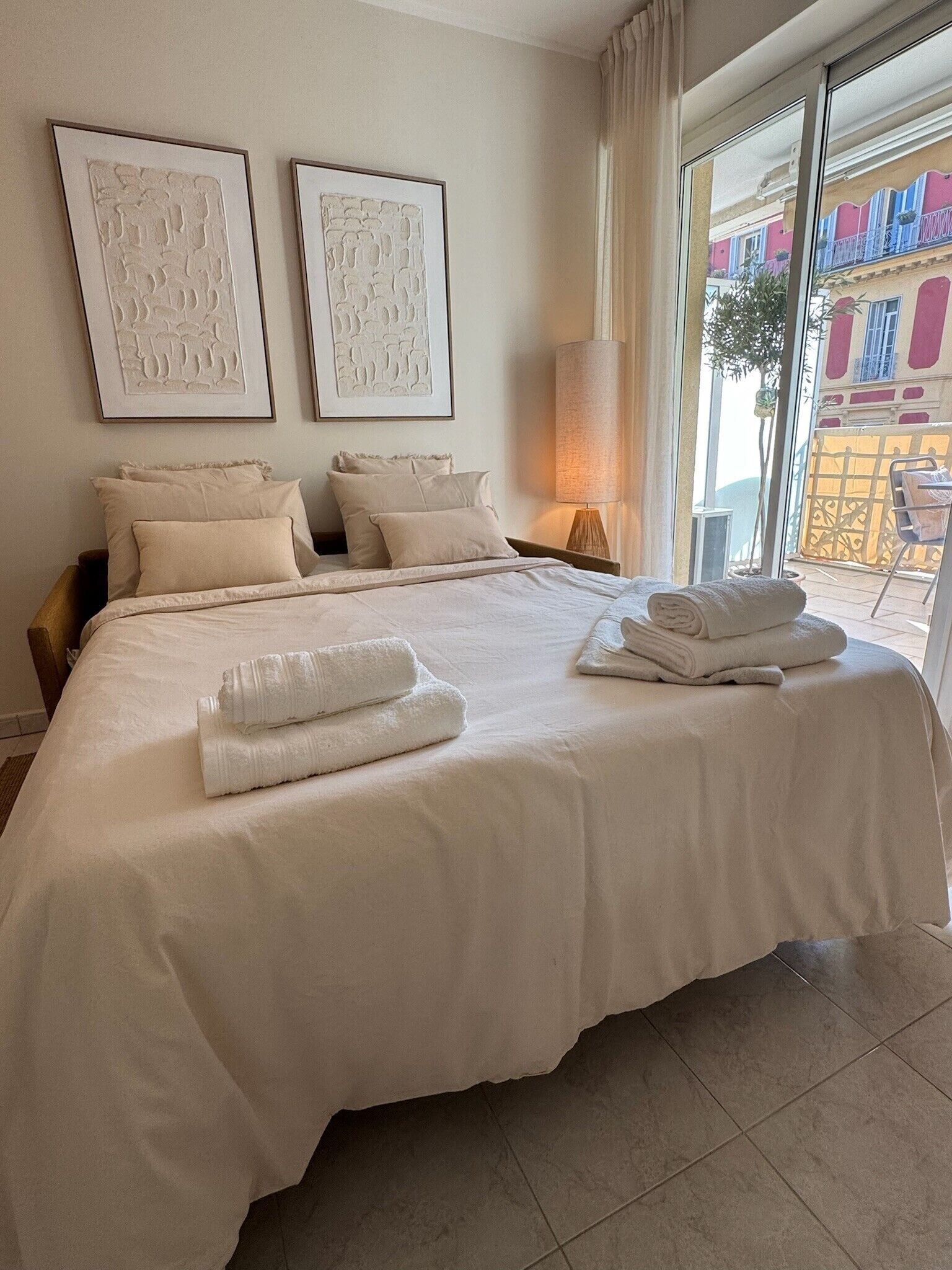 A hotel room with a large bed, two framed artworks on the wall, and a view of a building through the window.