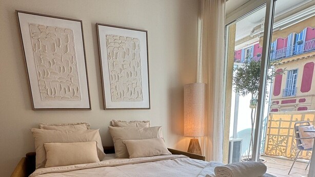 Ett hotellrum med en stor säng, två inramade konstverk på väggen och utsikt över en byggnad genom fönstret.
