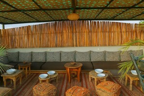 Un espacio al aire libre techado con un muro de bambú trenzado, asientos acolchados y mesas redondas de madera con cuencos.