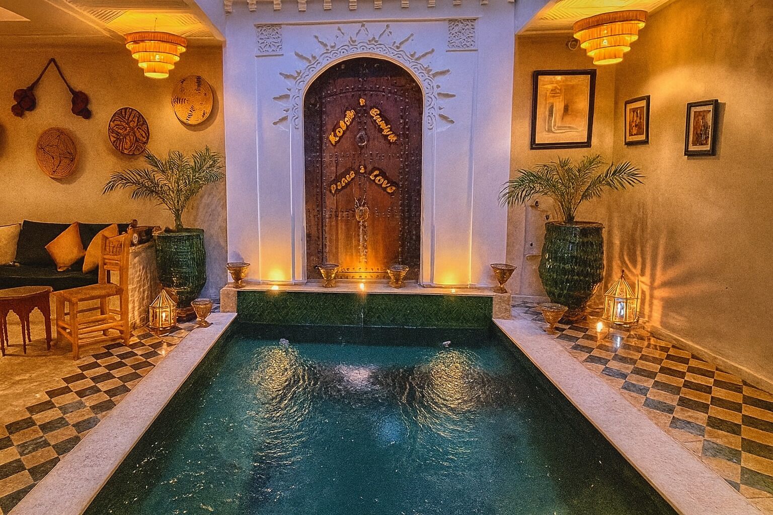 A pool area with a checkered floor, a wooden door with decorative carvings, and wall-mounted lights.