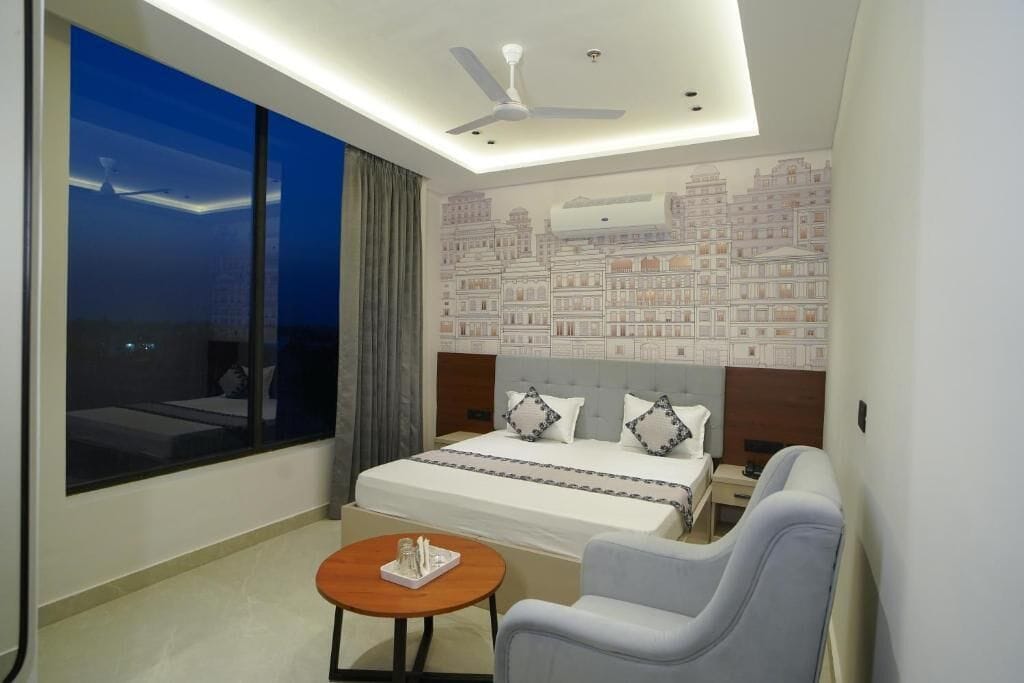 Una habitación de hotel con una cama grande, un escritorio, una silla, una mesa redonda, un ventilador de techo y vistas a edificios a través de la ventana.