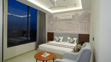 Una habitación de hotel con una cama grande, un escritorio, una silla, una mesa redonda, un ventilador de techo y vistas a edificios a través de la ventana.