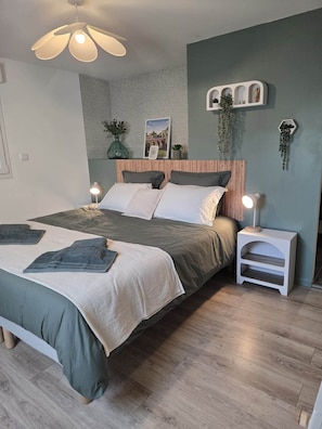 A modern bedroom with a wooden headboard, a large bed with multiple pillows, a bedside table with a lamp, and a wall-mounted shelf with plants. - Montr'oeil d'Opale (Écuires)