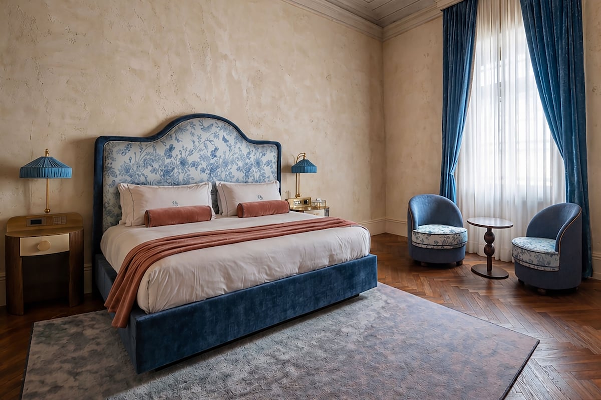Un dormitorio con una cama grande, dos mesitas de noche, un tocador, una silla, una pequeña mesa y una ventana con cortinas.