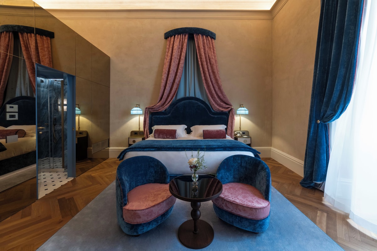 Una habitación de hotel con una cama grande, una zona de estar con dos sillones, una mesita y un espejo.