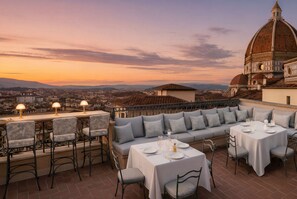 Un espacio de comedor al aire libre con mesas y sillas, con vistas a un paisaje urbano y un edificio histórico.