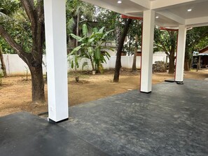 An outdoor covered area with white pillars and a concrete floor, surrounded by trees and a white wall.