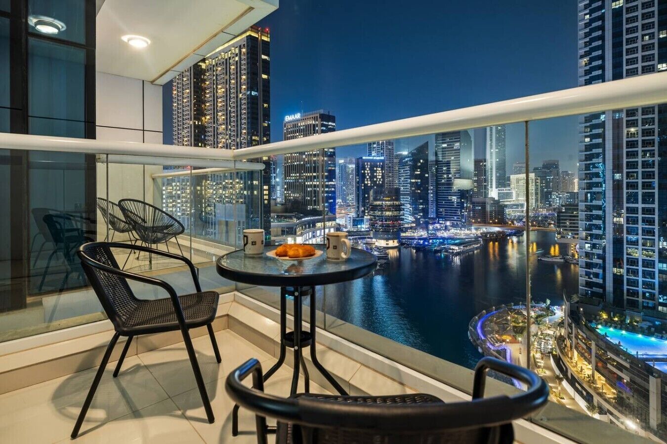 Un balcon avec vue sur la ville la nuit, comprenant une table avec des chaises et une tasse à café.
