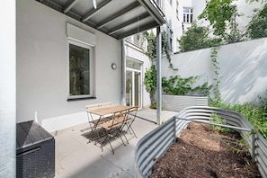 Eine Terrasse mit Tisch und Bänken, einem Mülleimer und einem Blumenbeet mit Pflanzen.