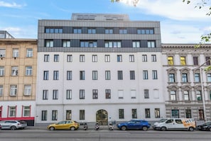 Ein mehrgeschossiges Gebäude mit moderner Fassade, einem zentralen Eingang und einem Parkplatz mit verschiedenen Fahrzeugen.