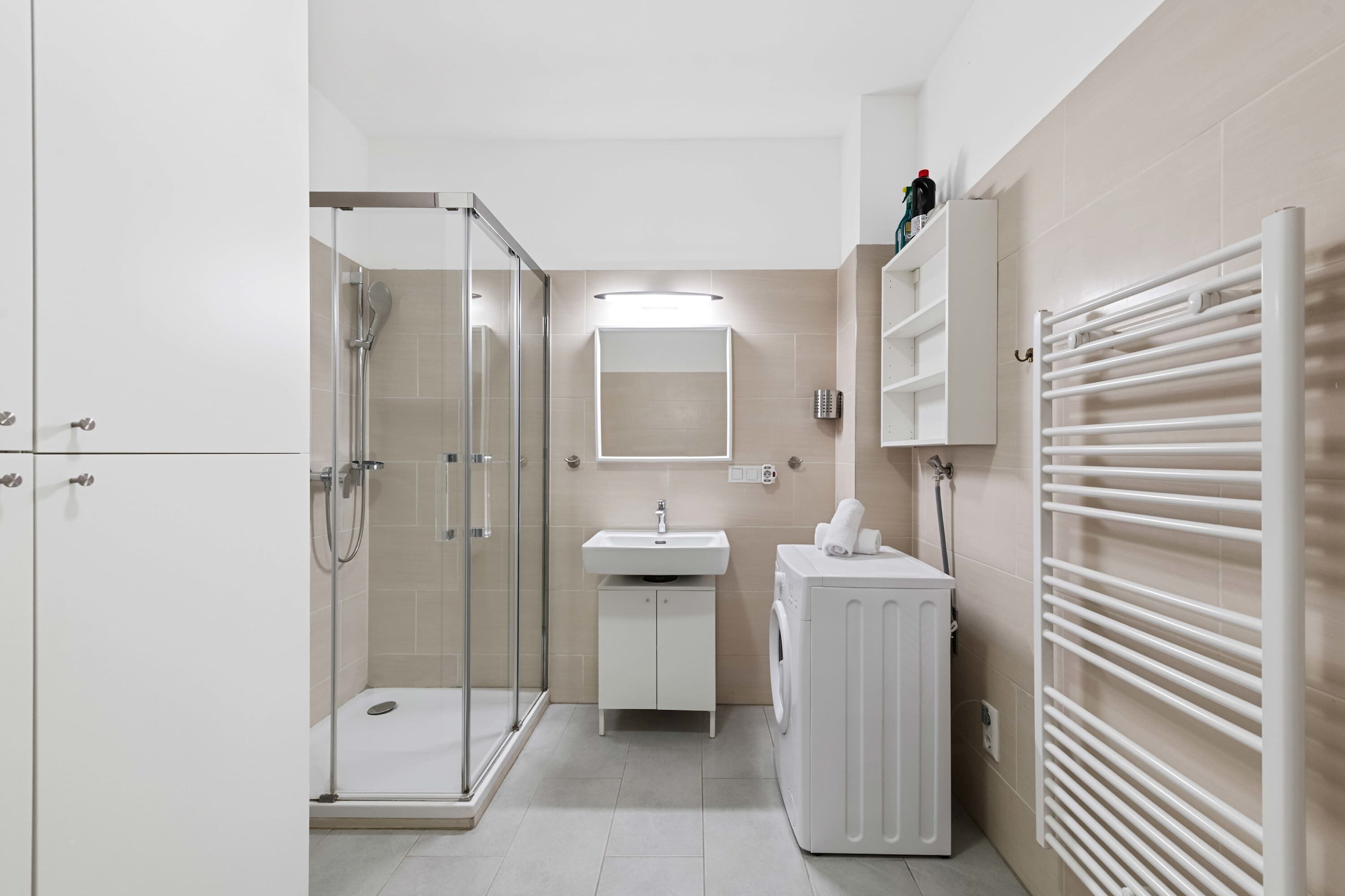 A modern bathroom with a glass-enclosed shower, a white sink, a built-in washing machine, and a towel rack.