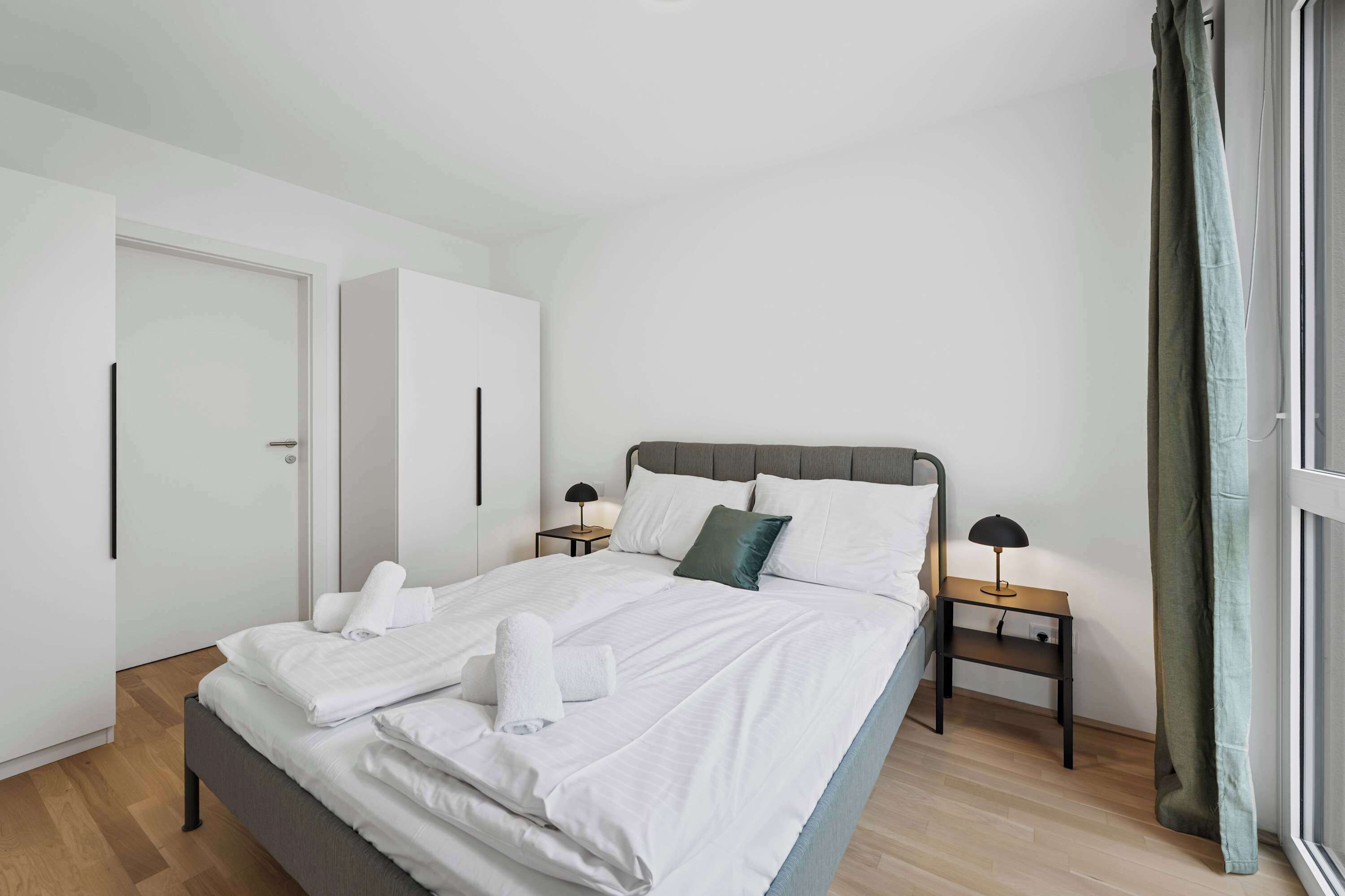 Ein modernes Hotelzimmer mit einem großen Bett, zwei Nachttischen mit Lampen, einem Kleiderschrank und Blick ins Freie.
