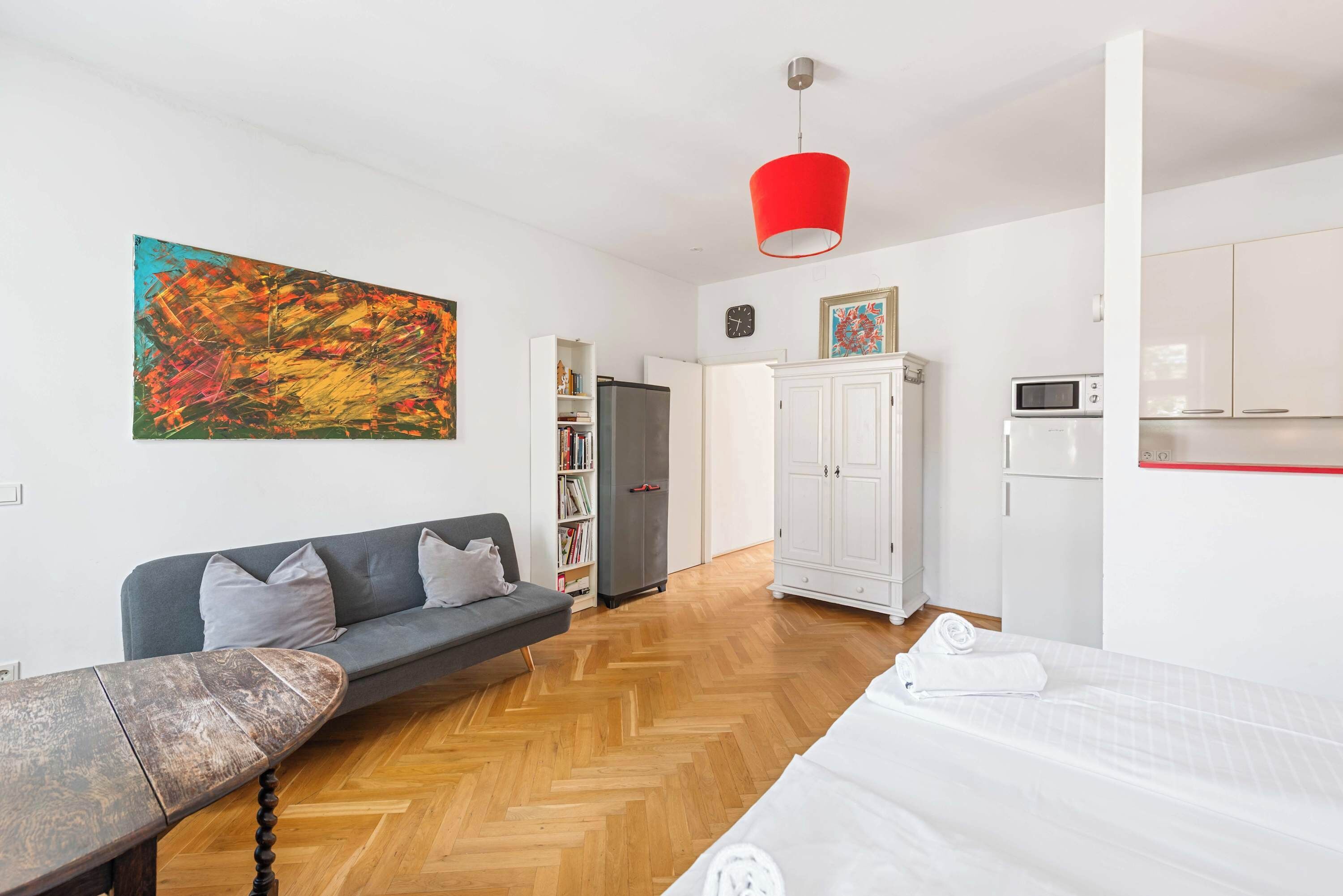 Ein modernes Schlafzimmer mit Holzdielen im Fischgrätmuster, einem grauen Sofa, einem weißen Bett, einer roten Pendelleuchte und einem weißen Kleiderschrank.