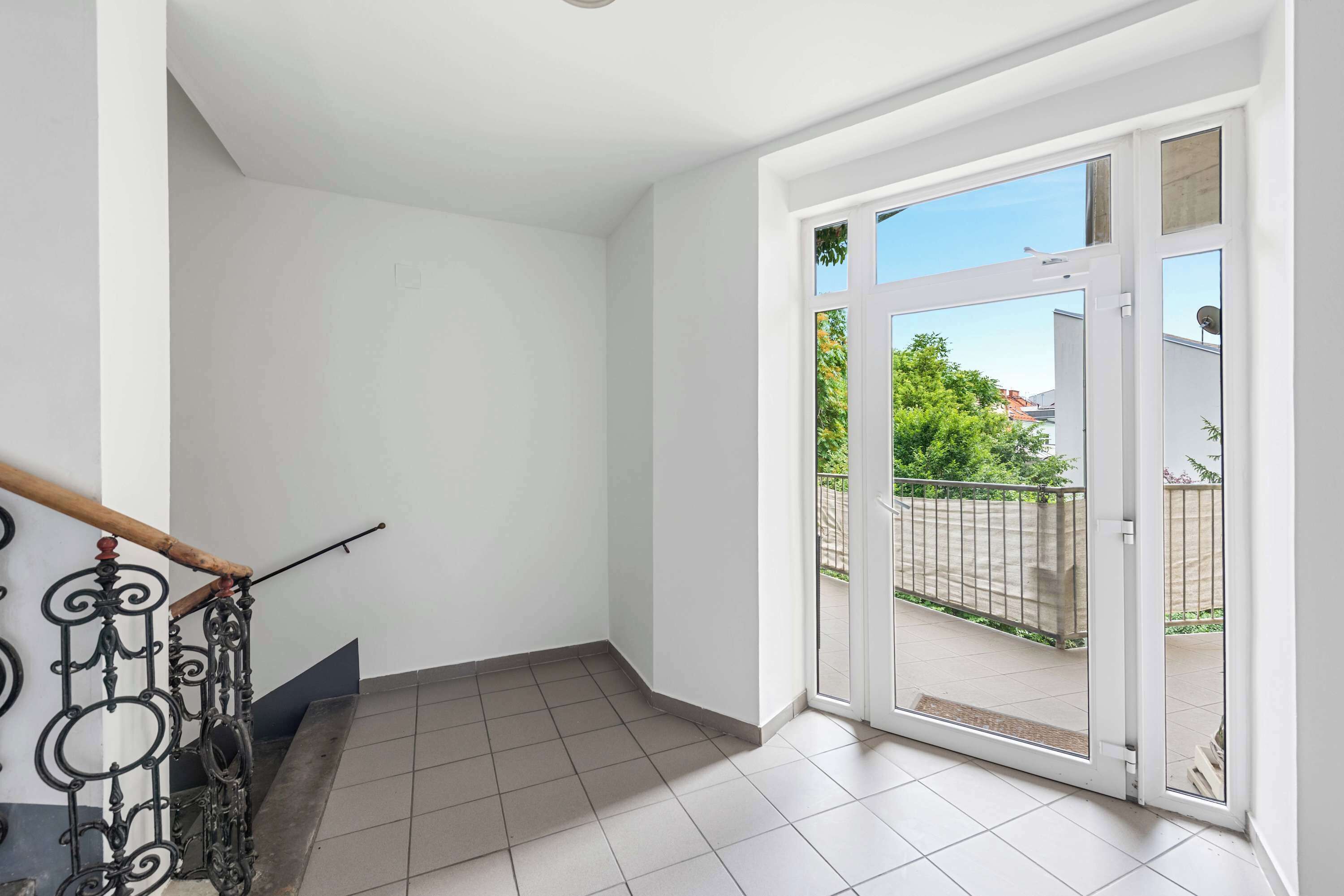 Una habitación con piso de baldosas, una escalera de madera con barandillas de metal decoradas y un amplio ventanal corredera que da a un balcón exterior.