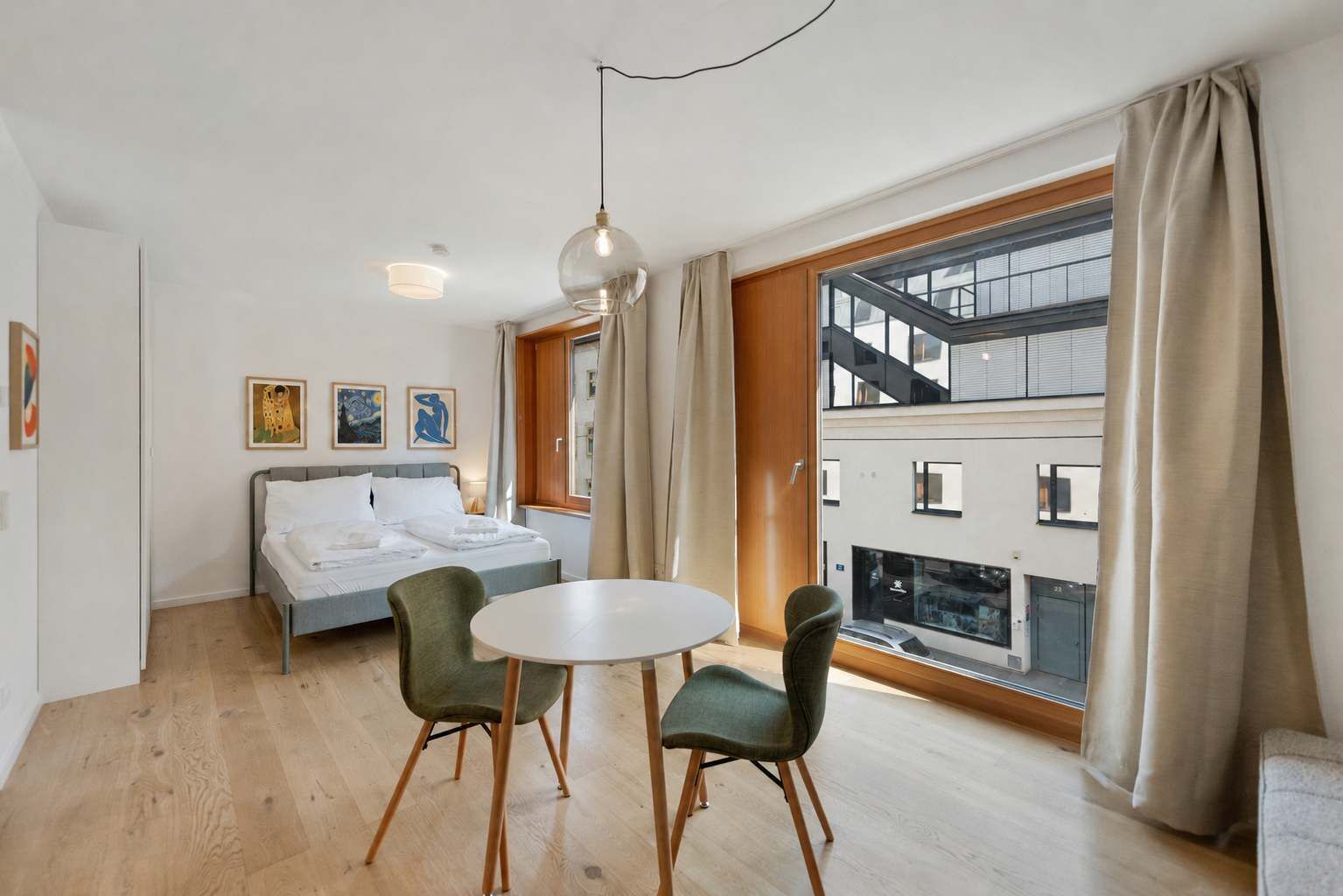 Ein modernes Hotelzimmer mit einem Bett, einem runden Tisch, zwei Stühlen, einem großen Fenster mit Vorhängen und Kunstwerken an der Wand.