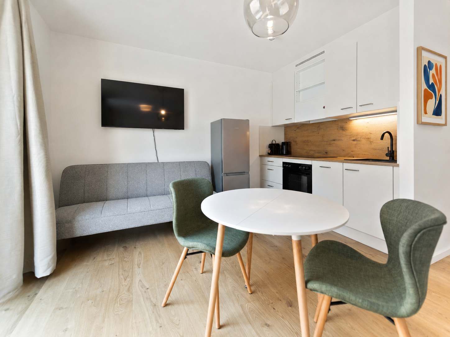 Un espacio habitable moderno con un sofá gris, una mesa redonda blanca y dos sillas verdes. Un televisor de pantalla plana está montado en la pared, y hay una zona de cocina con armarios blancos y una encimera de madera.