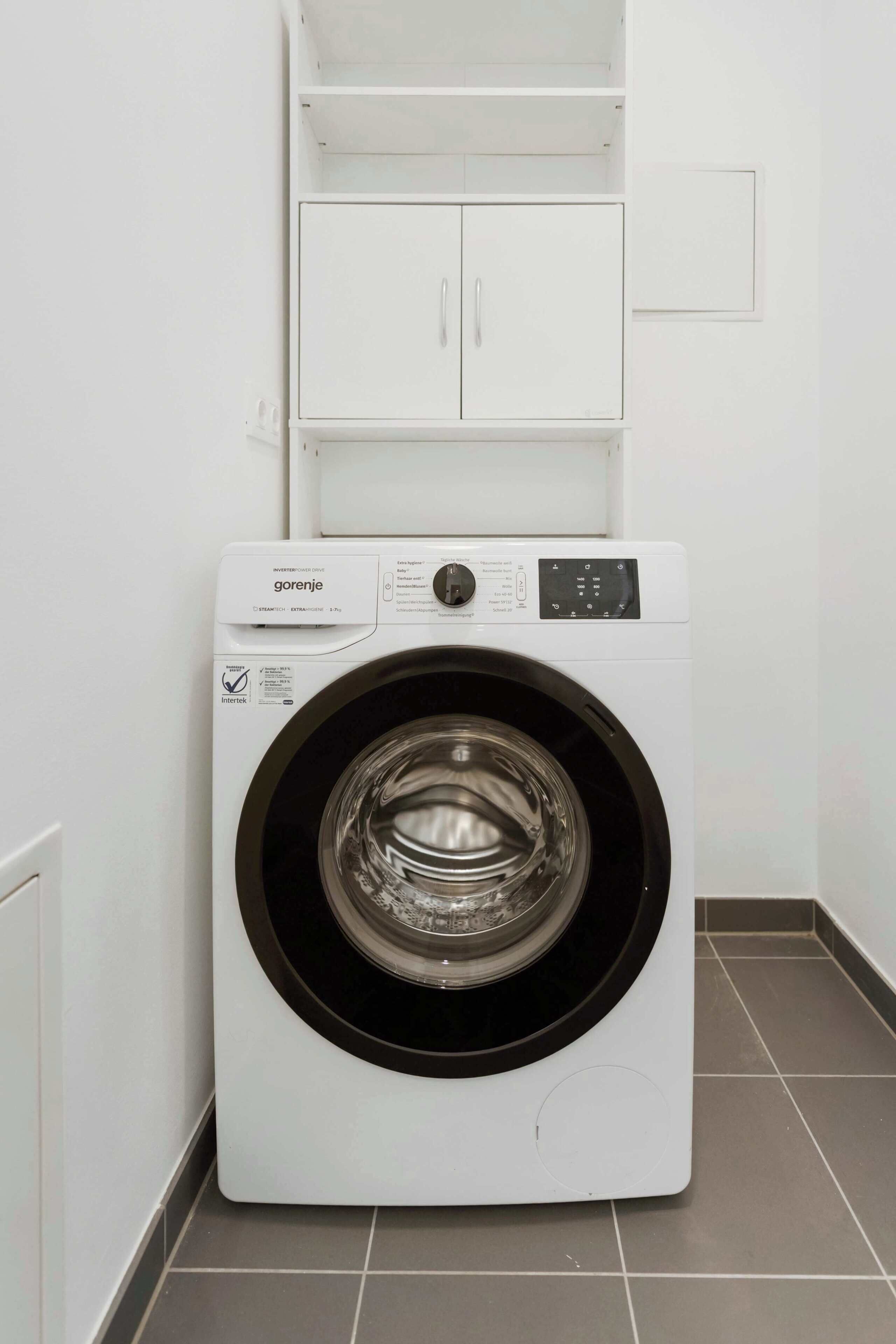 Une machine à laver blanche, dotée d’un panneau de commande et d’une porte transparente, est placée dans un coin, sur un sol carrelé.