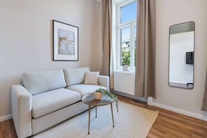 Ett modernt vardagsrum med en soffa, ett litet bord med en växt, en inramad tavla på väggen, ett fönster med gardiner och en spegel.