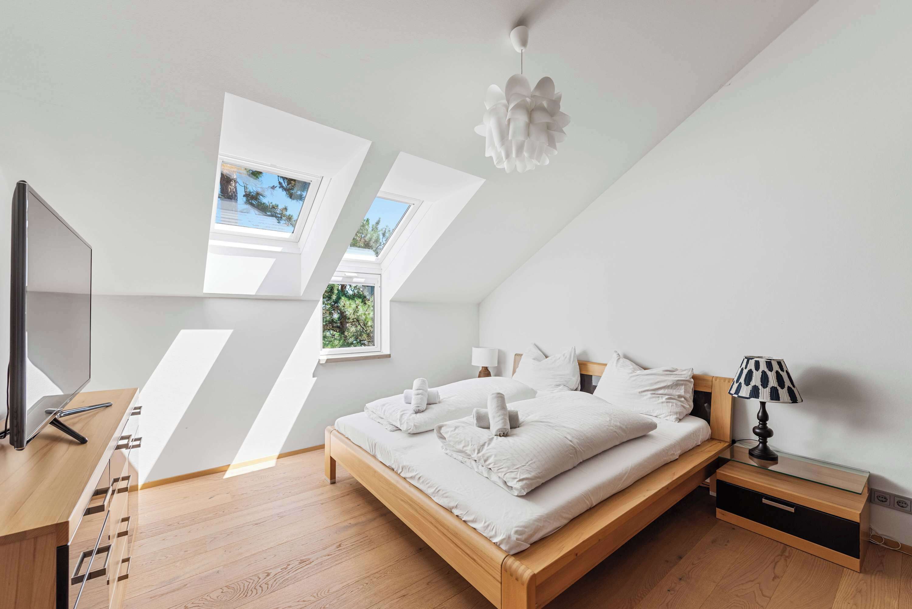 Ein modernes Schlafzimmer mit einem Holzbett, zwei Dachfenstern, einem Schreibtisch mit Flachbildfernseher und einem Nachttisch mit Lampe.