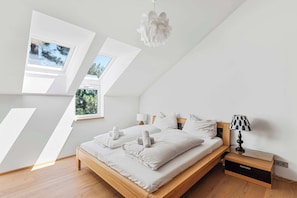 Een slaapkamer met een schuine kap, dakramen, een bed met witte lakens, een bedlampje en een nachtkastje.
