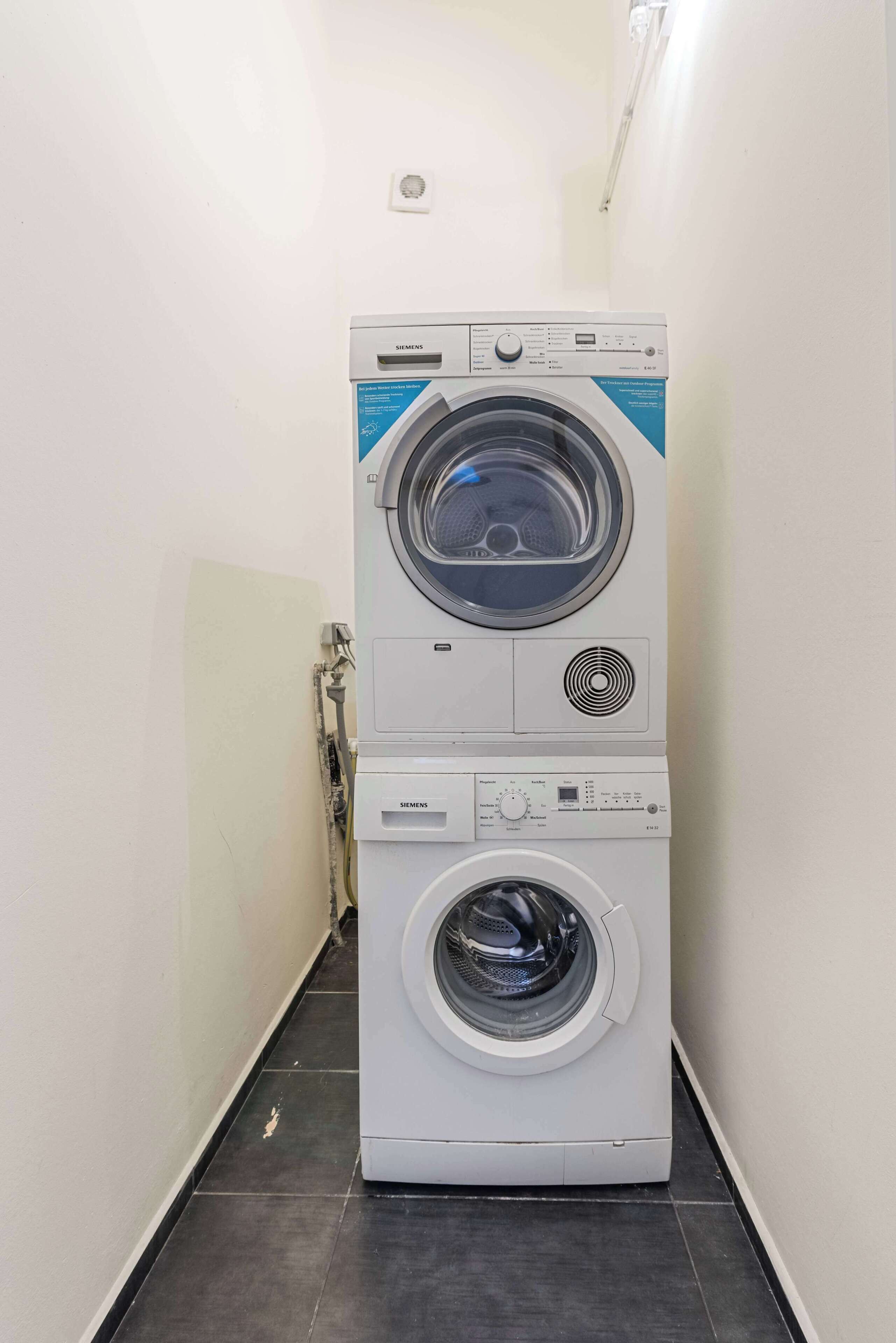Eine übereinander gestellte Waschmaschine und ein Trockner in einem schmalen Flur mit Fliesenboden.