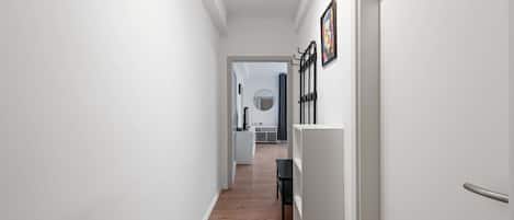 Un couloir aux murs blancs et au plancher en bois mène à une pièce où se trouvent un lit, un miroir et un présentoir en métal noir.
