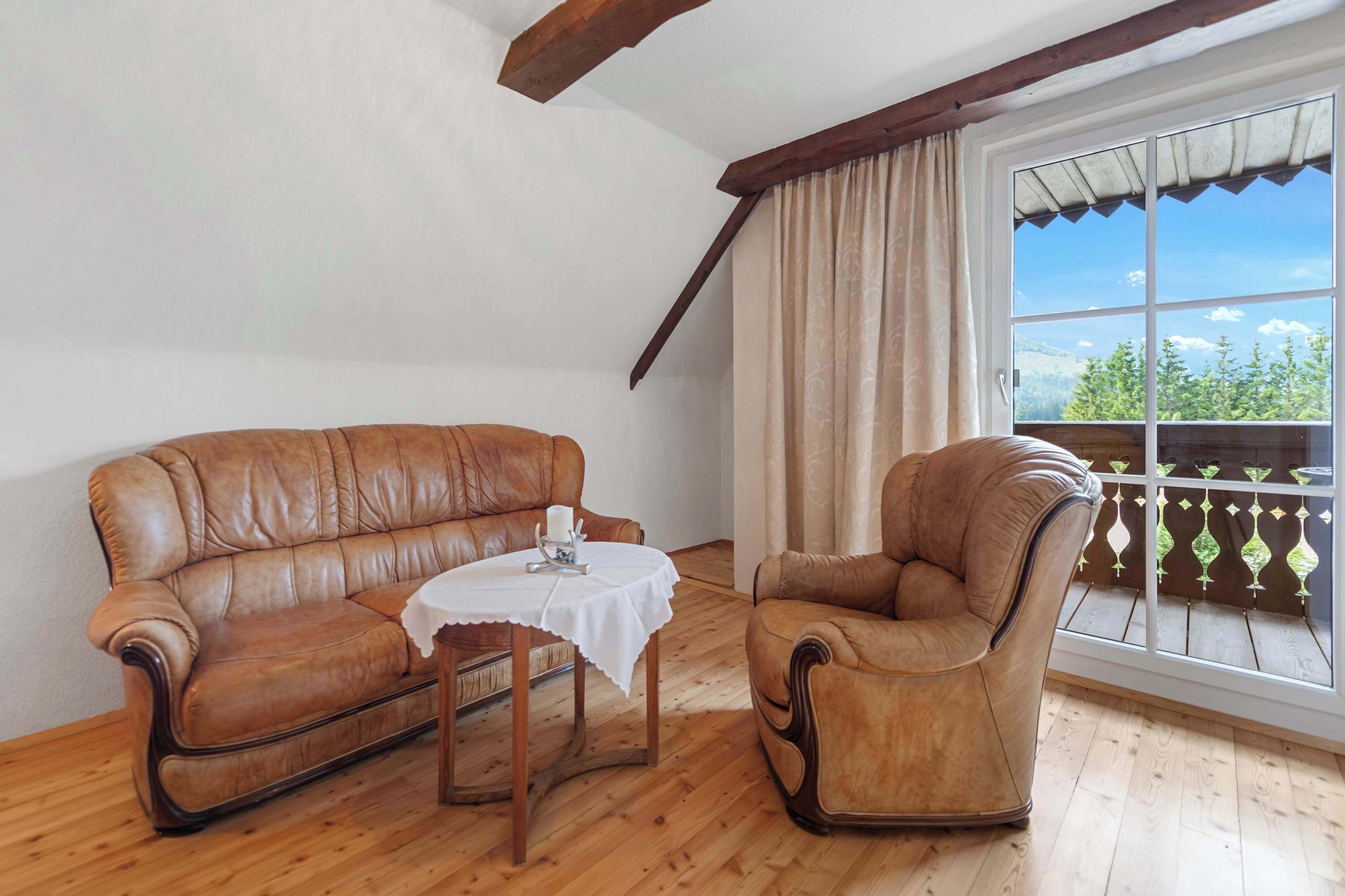 A room with a wooden floor, a brown leather sofa, a brown leather armchair, and a small round table with a white tablecloth.