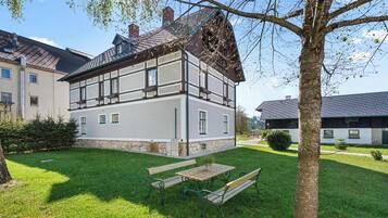 A two-story building with a sloped roof, a stone foundation, and a small garden with a bench and a table.