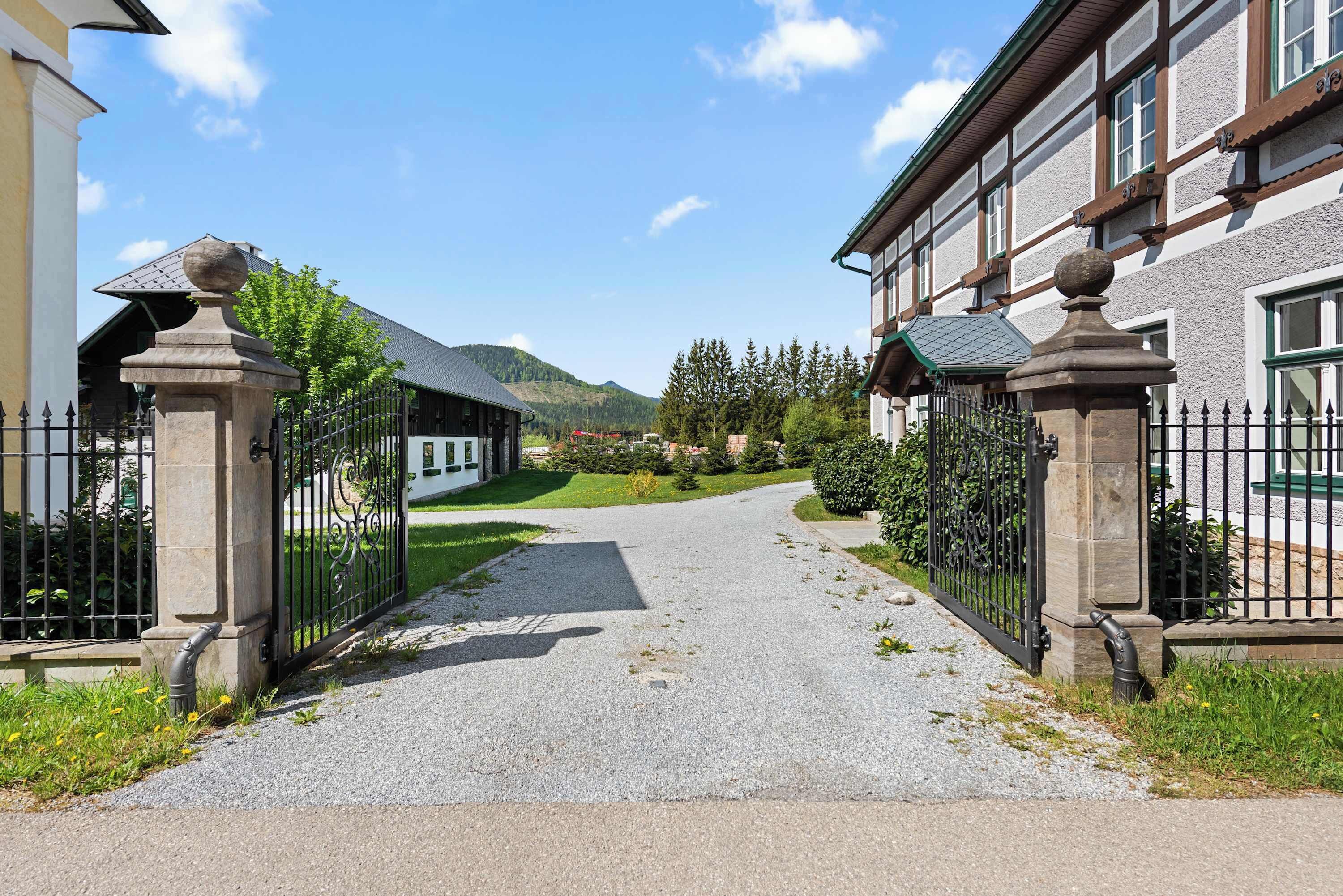 Ein befestigter Zugangsweg führt zu einem Gebäude im traditionellen Baustil, umgeben von Grünflächen und einer Bergkulisse.