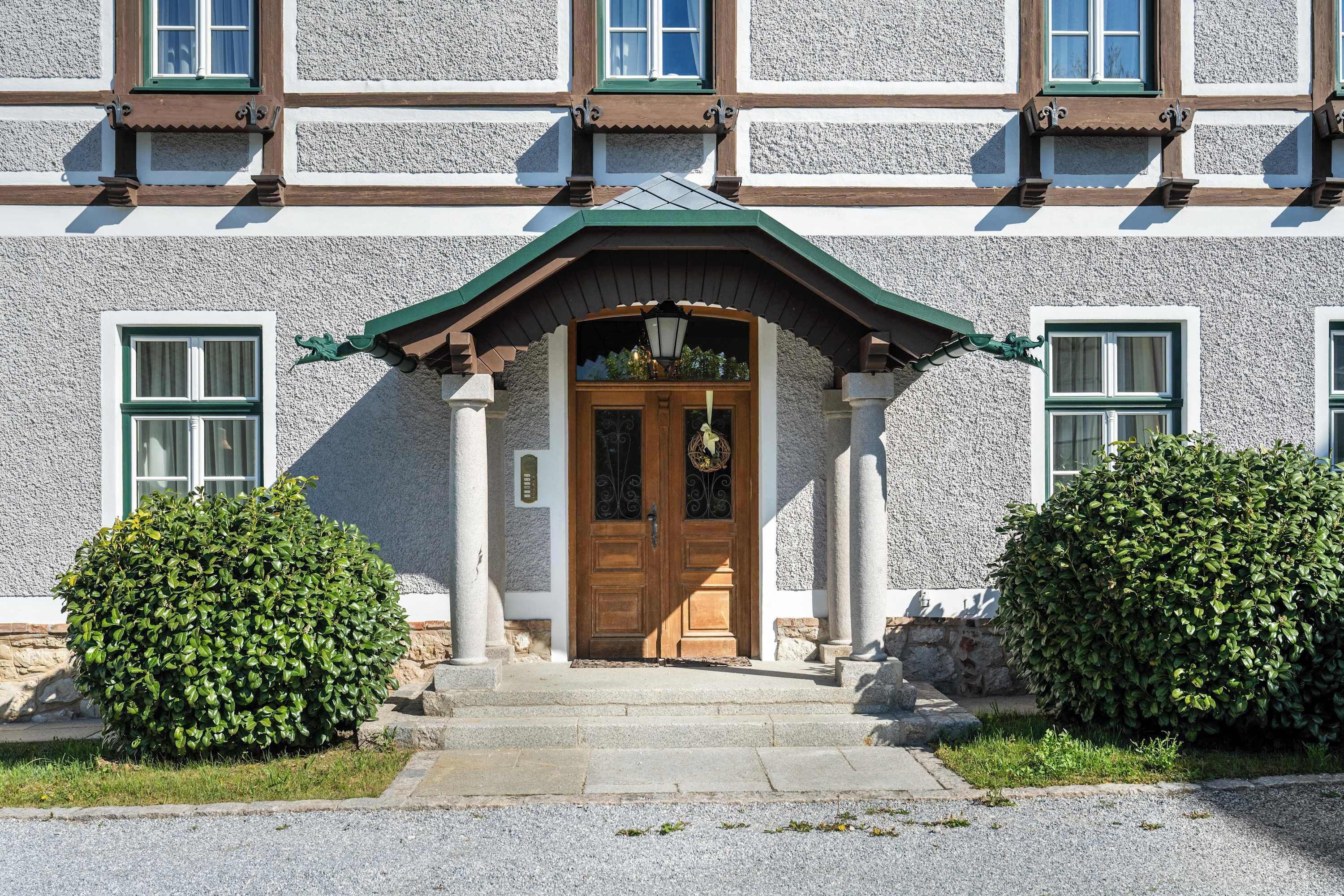 A building with a traditional architectural style, featuring a symmetrical facade, a central entrance with a wooden door, and a green roof.