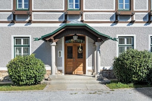 A building with a traditional architectural style, featuring a symmetrical facade, a central entrance with a wooden door, and a green roof.
