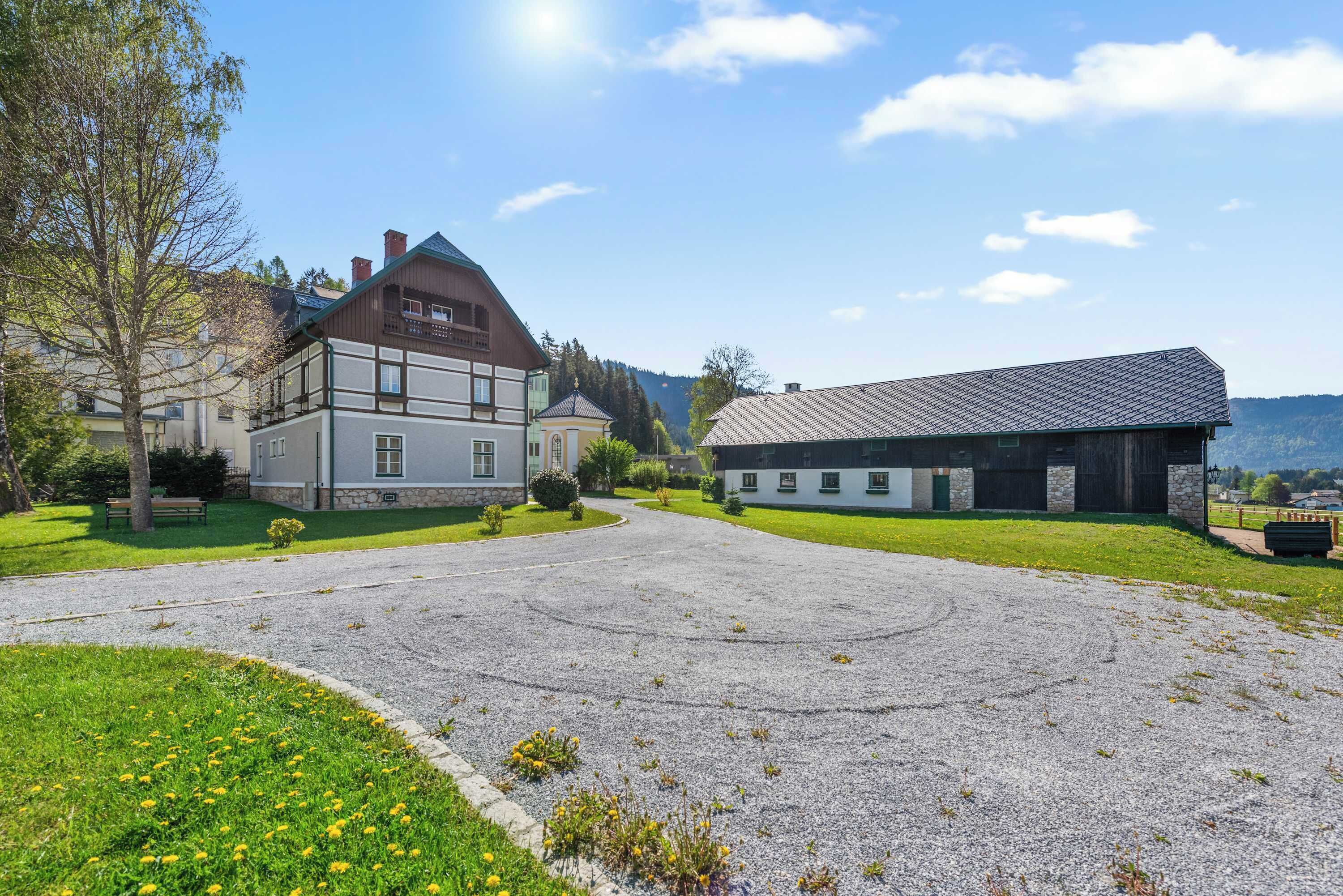 A building with a traditional architectural style, a paved driveway, and a grassy area with a bench.