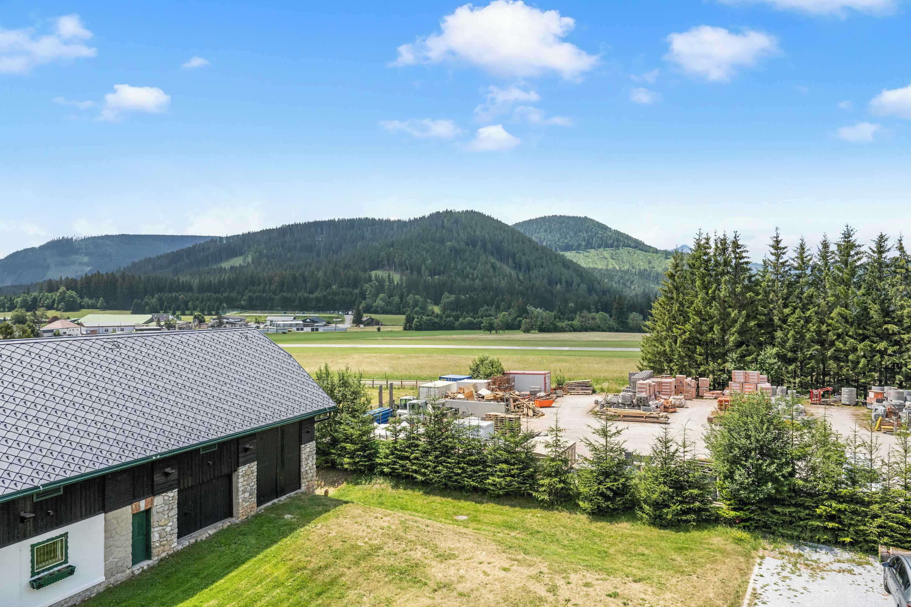 A rural building with a sloped roof, surrounded by a grassy area and a forested mountain backdrop.