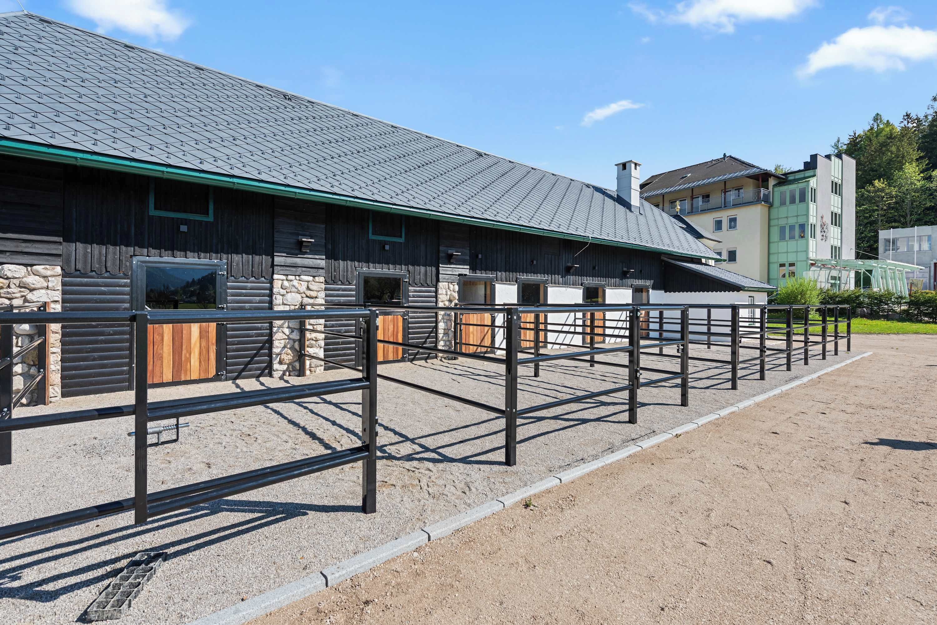 A stable with a metal fence, stone walls, and a green roof.