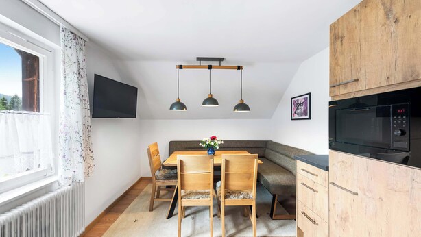 A modern kitchen with wooden cabinets, a dining table with chairs, pendant lights, and a large window with curtains.
