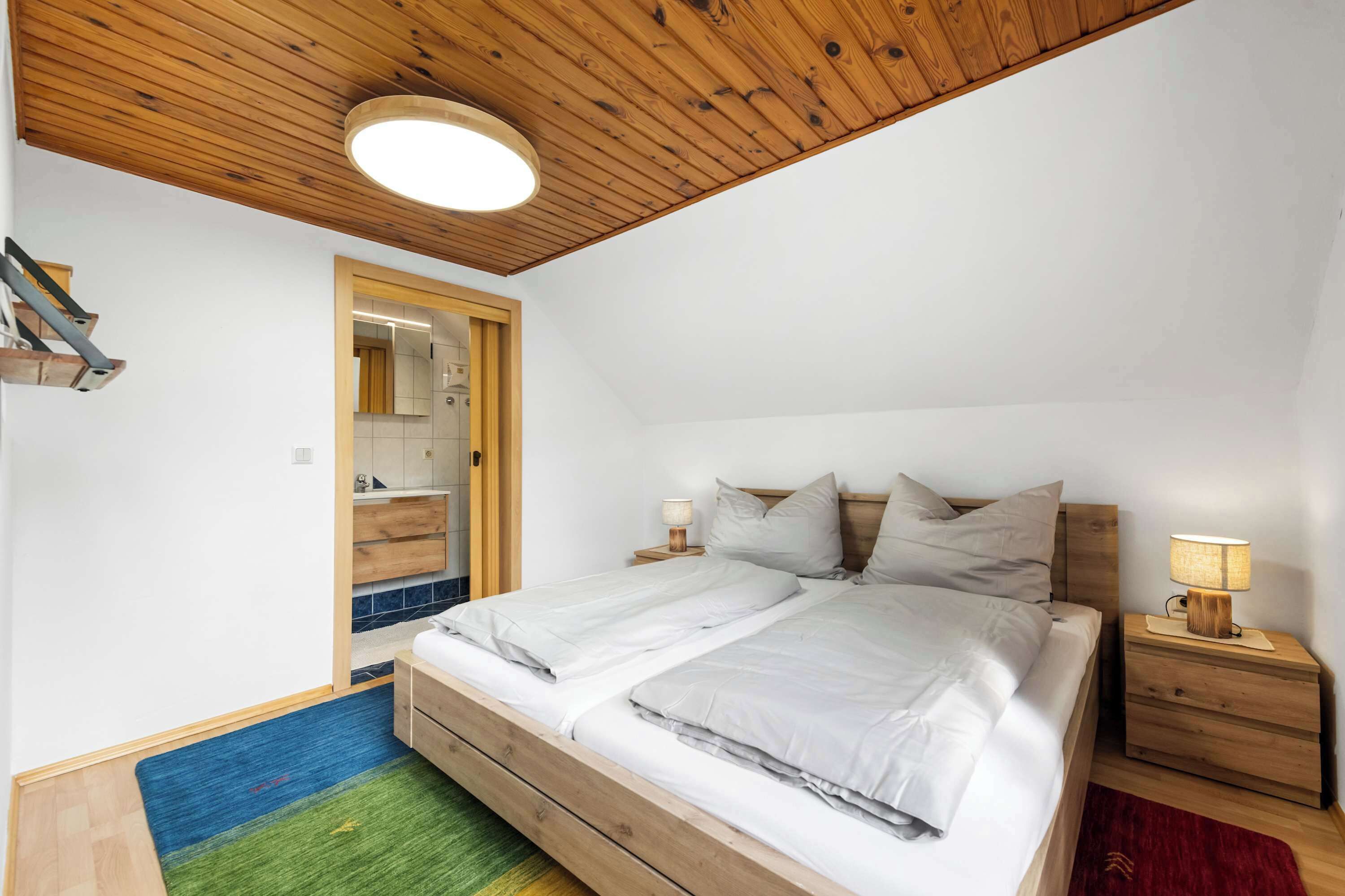 Una camera da letto con soffitto in legno, un letto con biancheria da letto bianca, un comodino con una lampada e un bagno visibile attraverso una porta aperta.