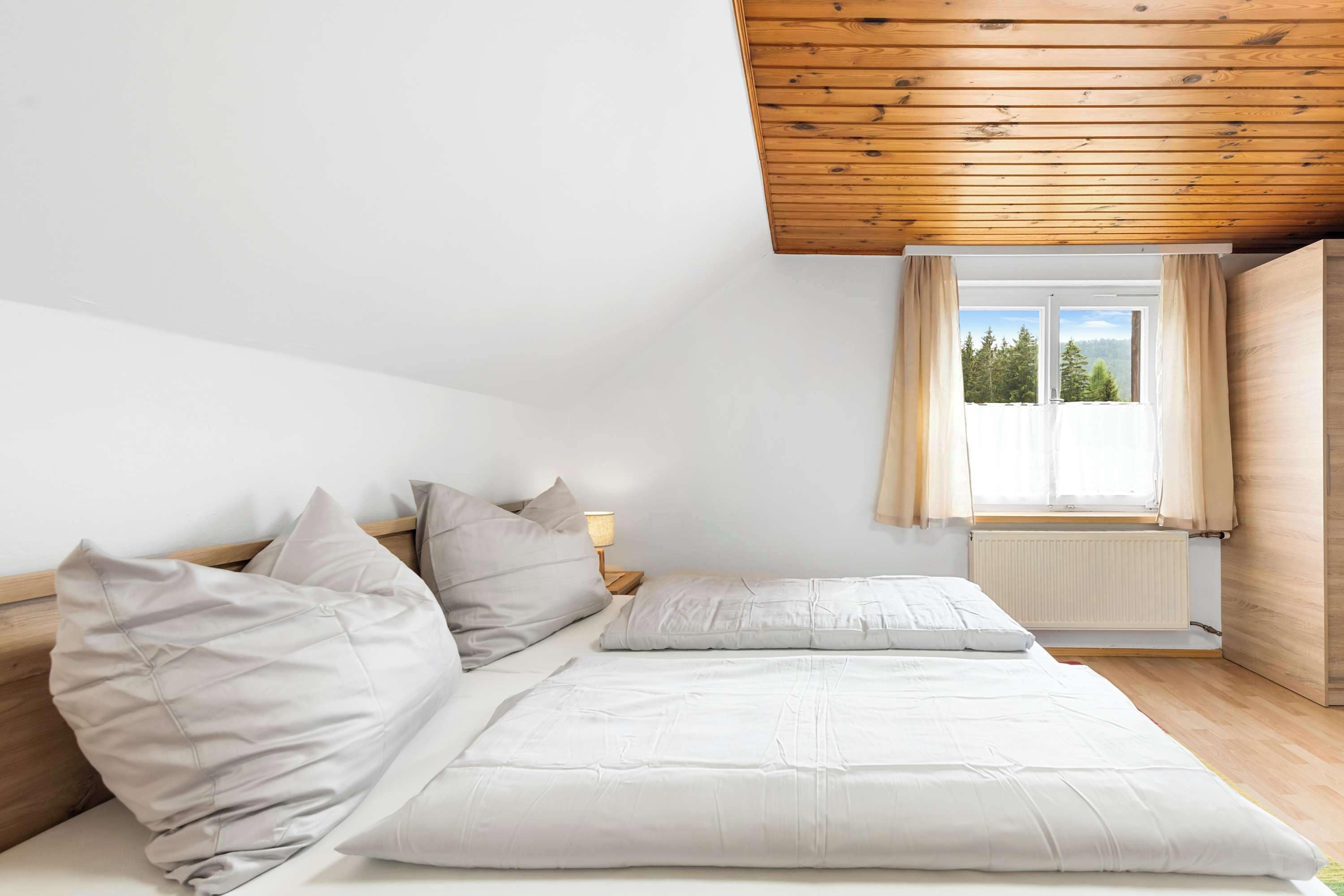Una camera da letto con un letto, cuscini, un comodino, una finestra con tende e vista sugli alberi.