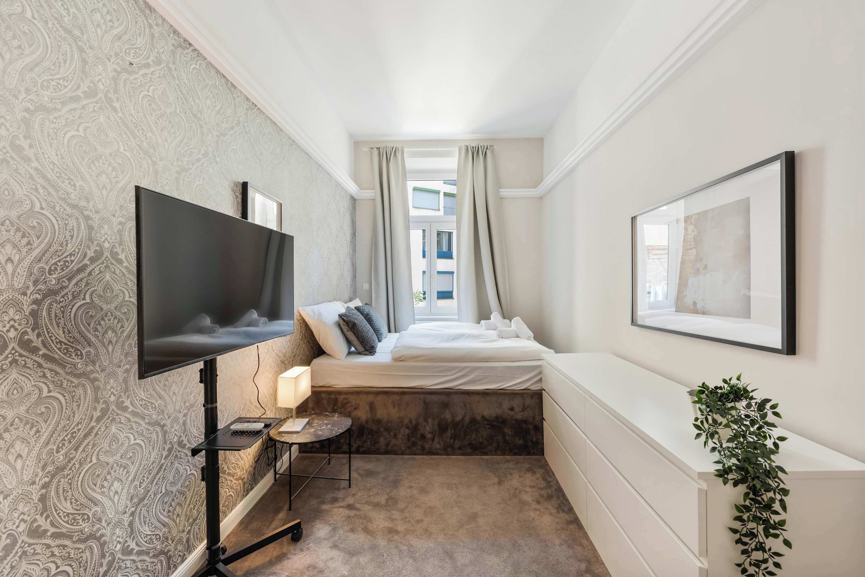 A modern hotel room with a large flat-screen TV mounted on the wall, a bed with white linens, a small bedside table with a lamp, and a potted plant.