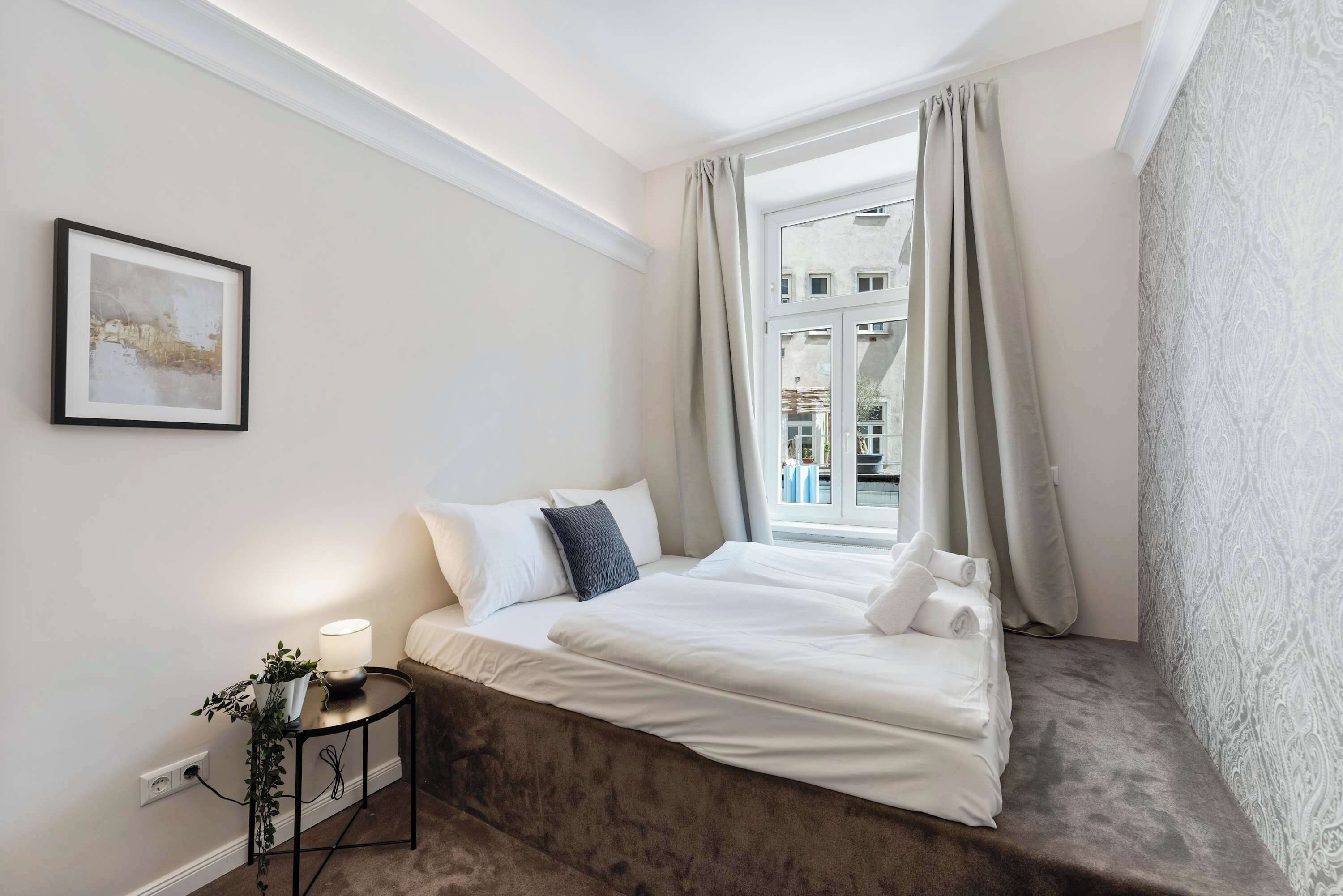 A hotel room with a bed, a bedside table with a lamp, a power outlet, and a window with curtains.
