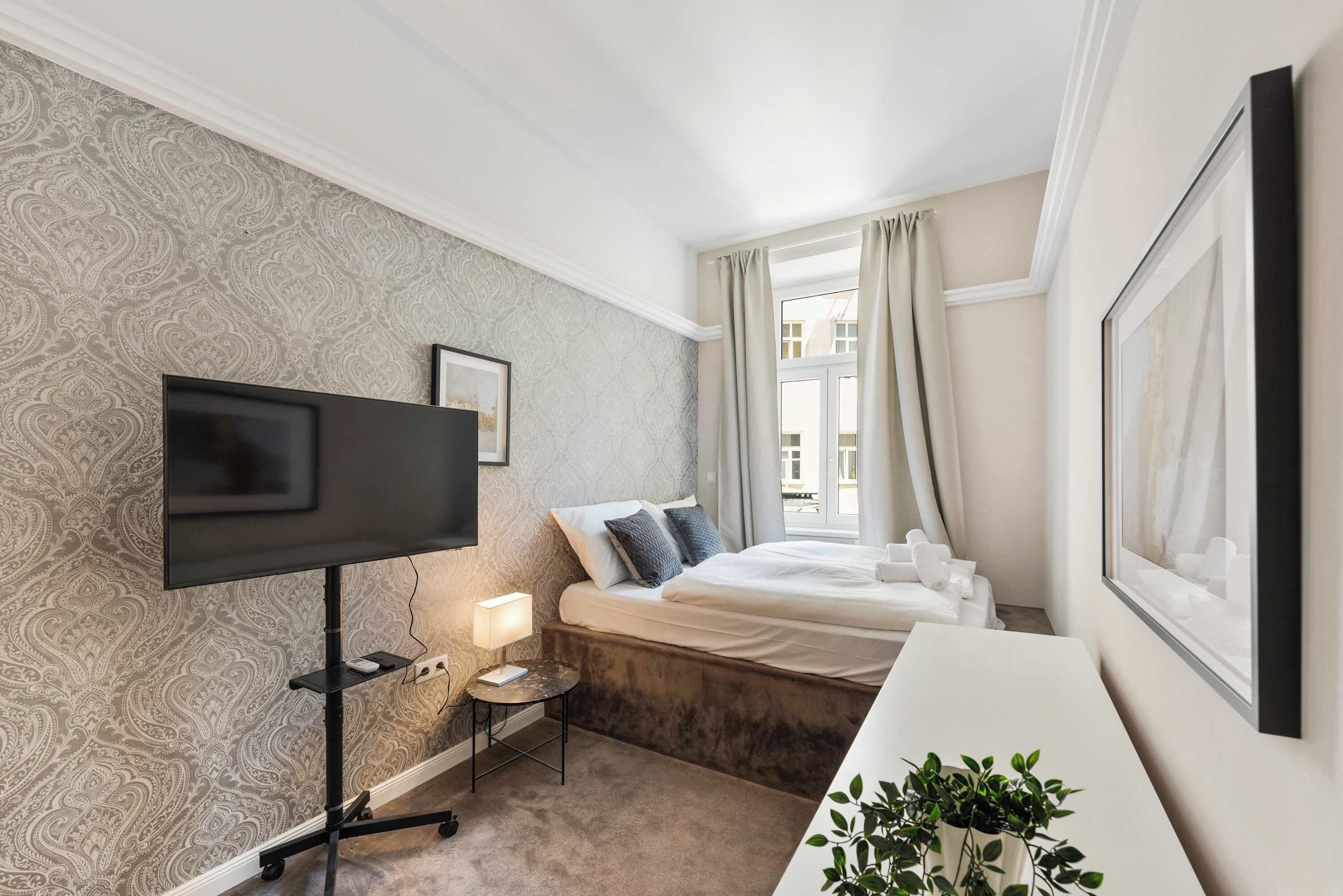 Een hotelkamer met een grote flatscreen-tv, een nachtkastje met lamp, een bed met kussens en een raam met gordijnen.