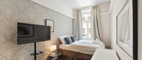 Una camera d'albergo con una grande TV a schermo piatto, un comodino con una lampada, un letto con cuscini e una finestra con tende.