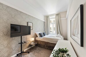 A hotel room with a large flat-screen TV, a bedside table with a lamp, a bed with pillows, and a window with curtains. - Kaiser 2BR | Metro & Tram | Nespresso | Central (Wien)