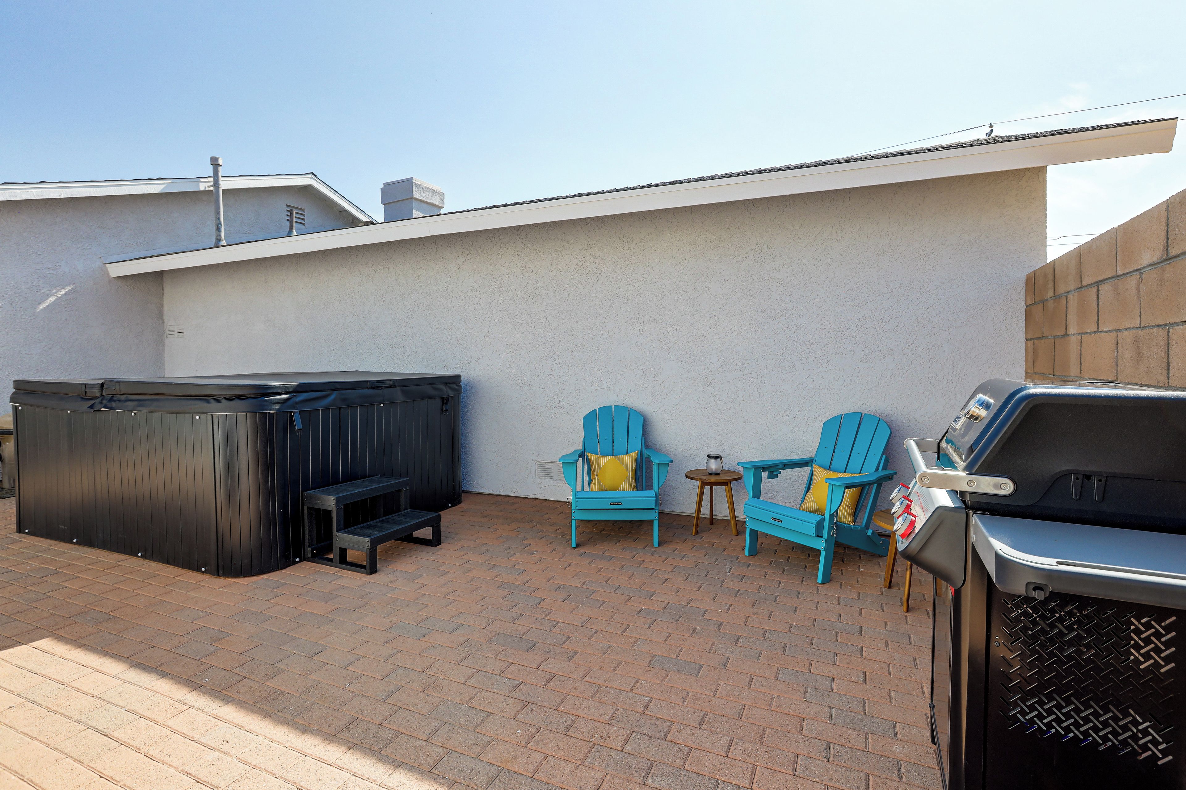 Un patio con jacuzzi, dos sillas Adirondack, una parrilla y piso de ladrillo.
