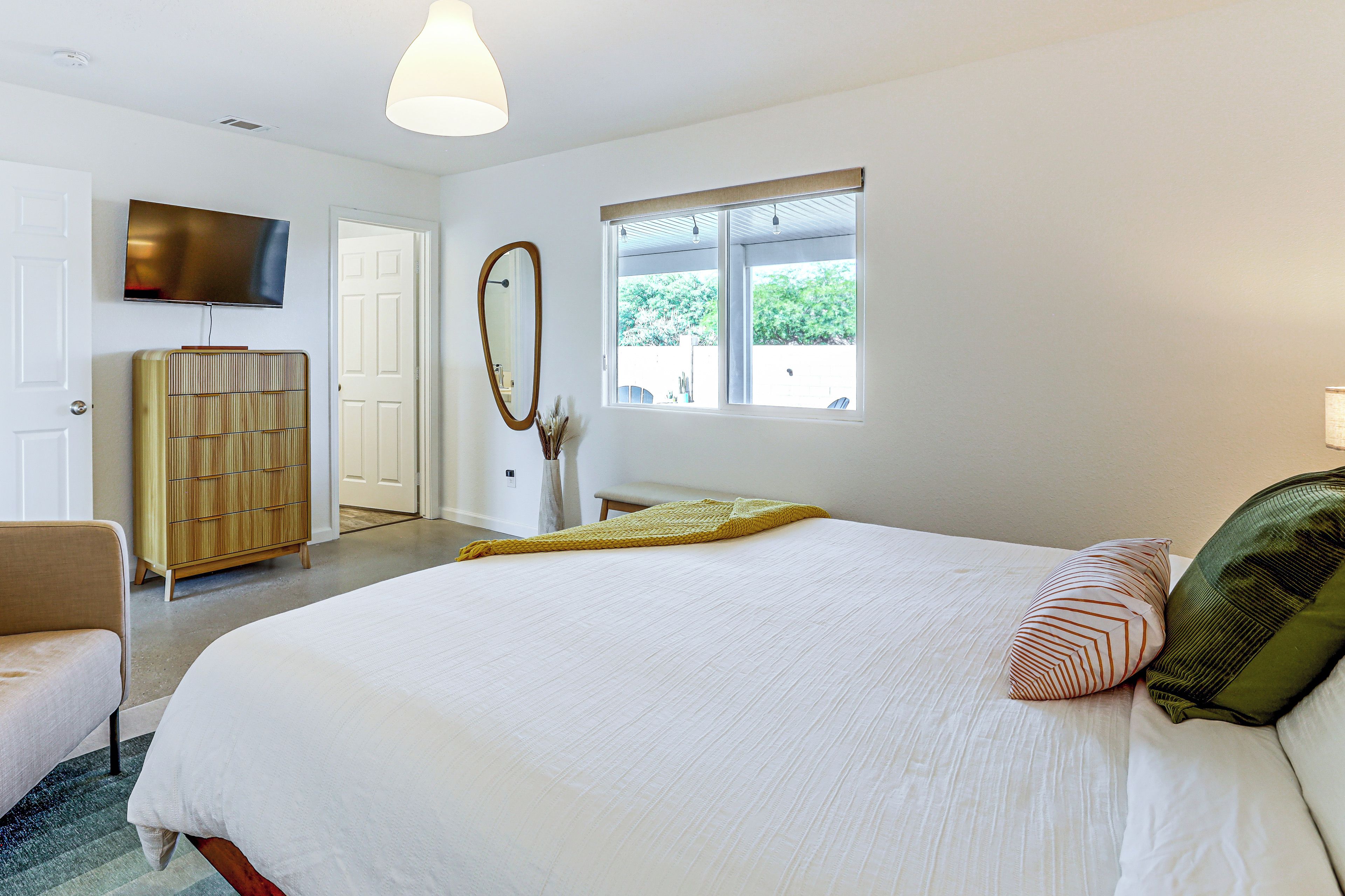 A bedroom with a large bed, a television mounted on the wall, a window with a view of trees, and a door leading to another room.
